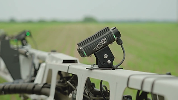 Black camera on equipment in a green field, mounted on a metal frame. Device has a simple design. Calm rural environment in the background.