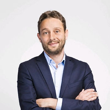 A man in a navy suit with a light blue shirt stands with arms crossed against a white background, smiling confidently.